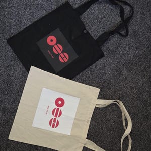 May include: Two tote bags, one black and one beige, are displayed on a gray surface. Each bag features a rectangular patch with a red graphic design and the text "2016.05.20". The bags have long handles.