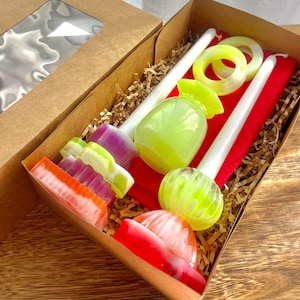 May include: A gift box filled with colorful, decorative soaps and candles. The box contains a variety of soaps in shades of red, orange, purple, and yellow. There are also two white candles, a green vase, and two yellow rings. The box is lined with red fabric and brown paper.