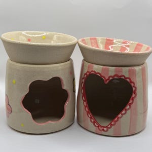 May include: Two ceramic wax warmers. One is beige with a flower cutout and painted flowers. The other is pink and white striped with a heart cutout. Both have a matching beige dish on top with decorative accents.