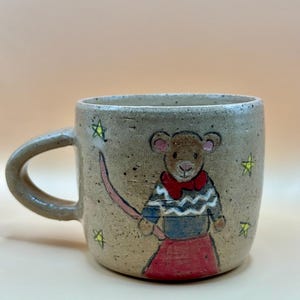 May include: A hand-painted ceramic mug with a mouse design. The mug has a speckled, earthy tone and features a mouse wearing a sweater and skirt, with a pink tail. Yellow stars are scattered around the mouse. The mug has a handle.