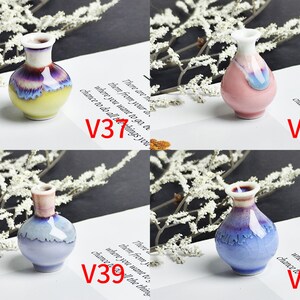 Op de afbeelding: Vier miniatuur keramische vazen met de labels V31, V38, V39 en V40. V31 is geel en paars, V38 is roze en wit, V39 is lichtblauw en wit, en V40 is blauw en paars met een craquel&eacute; effect. Elke vaas heeft een smalle hals en een ronde vorm.