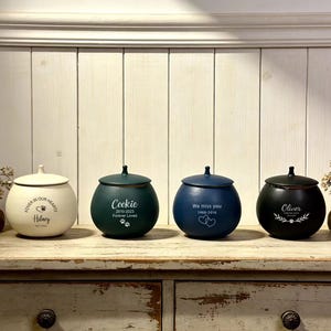 May include: Four ceramic pet urns in cream, dark green, navy blue, and black, each with a lid and personalized text. The urns are displayed on a wooden surface with decorative elements. The urns are for pets.