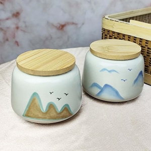 May include: Two white ceramic jars with bamboo lids. Each jar features a painted landscape design with mountains in shades of blue and brown, and small black bird silhouettes. The jars are on a light pink surface.