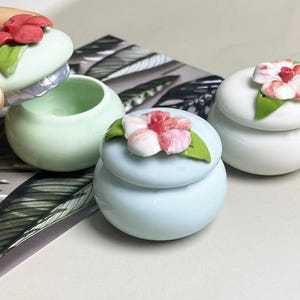 May include: Three small, round ceramic trinket boxes in shades of mint green, light blue, and white. Each box is topped with a decorative flower in shades of red, pink, and white with green leaves. One lid is open, revealing the inside of the box.