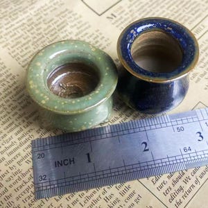 May include: Two ceramic ear gauges. One is light green with white speckles, the other is navy blue with a gold rim. A ruler shows the gauges are approximately 1 inch in diameter.