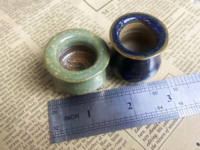 May include: Two ceramic ear gauges. One is light green with white speckles, the other is navy blue with a gold rim. A ruler shows the gauges are approximately 1 inch in diameter.