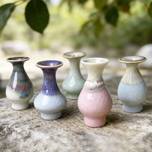 May include: Five small ceramic vases with a crackle glaze finish, each featuring a distinct color palette including pink, purple, blue, green, and white, with a bulbous body, a narrow neck, and a flared rim, arranged in a cluster.