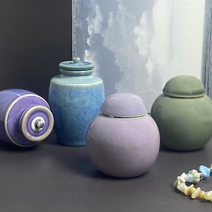 Peut inclure: Quatre pots en céramique de couleurs violet, bleu et vert, avec couvercles assortis. Un pot est ovale, les autres sont ronds. Un bracelet de perles avec un charme en forme d'étoile de mer est au premier plan.