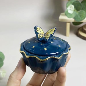 May include: A navy blue ceramic trinket box with a butterfly lid. The box has a scalloped edge with gold trim and gold polka dots. The butterfly is yellow and black.