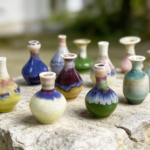 May include: A collection of small, decorative ceramic vases in various colors, including blue, green, yellow, and red. The vases have unique shapes and painted designs, displayed on a stone surface. The collection showcases a variety of artistic styles.