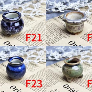 May include: Four small ceramic pots in various colors: dark blue with white swirls, beige with purple accents, cobalt blue, and green with brown trim. Each pot has a unique shape and sits on three small feet. The pots are displayed on a newspaper background.