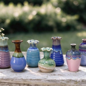May include: A collection of seven small ceramic vases with varied shapes and glossy glaze finishes in shades of purple, blue, green, and pink. Each vase features a distinct neck design, including ruffled, flared, and narrow openings.
