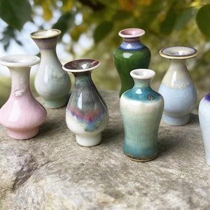 May include: A collection of small, decorative ceramic vases in various colors, including pink, green, blue, and white. Each vase has a unique shape and glaze, with a glossy finish. The vases are arranged on a stone surface.