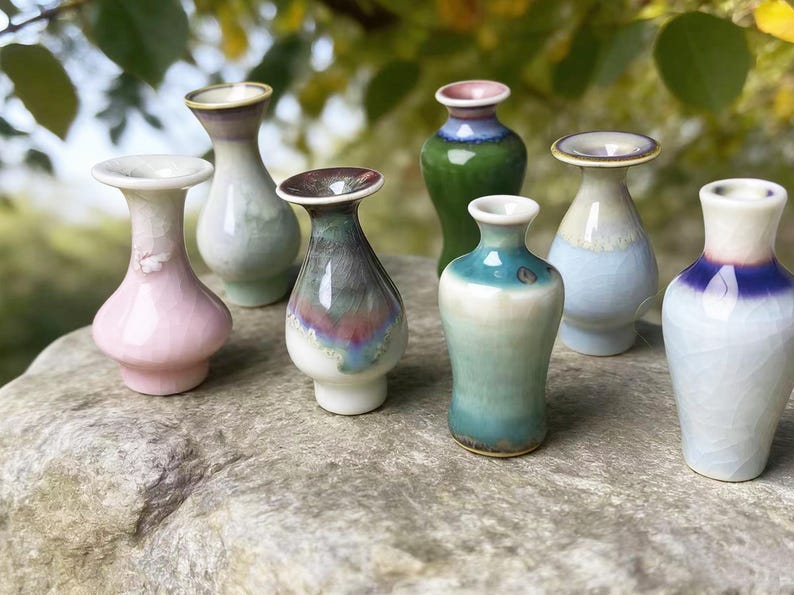 May include: A collection of seven small ceramic vases with flared rims and smooth, glossy glazes in various colors including pink, green, blue, and white. Each vase features a unique reactive glaze finish with subtle crackle patterns and gradients.