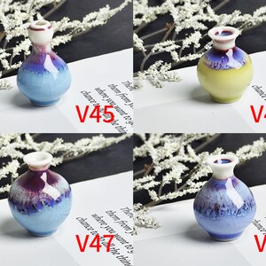 Op de afbeelding: Vier miniatuur keramische vazen gelabeld V45, V46, V47 en V48. V45 is lichtblauw met een donkerpaarse rand. V46 is geel met een donkerpaarse rand. V47 en V48 zijn lichtblauw met donkerpaarse glazuurdruppels, elk met een witte hals en rand.