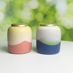 May include: Two ceramic jars with gold lids. One is yellow, white, and coral. The other is white, green, and blue. The jars have a rounded shape, possibly for storage or decorative use.