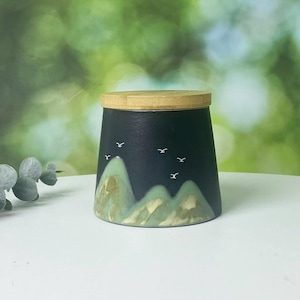 Peut inclure: Un pot en céramique noir avec un couvercle en bambou. Le pot présente un paysage avec des montagnes vertes et des oiseaux blancs. Le pot est sur une surface blanche avec un arrière-plan vert flou.