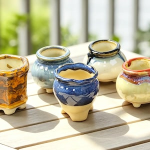 May include: Five small ceramic pots with varied glazes and shapes. One is square and amber-coloured, while the others are rounded with blue, white, and cream tones. Each pot features a wide opening, a thick rim, and three small feet at the base.