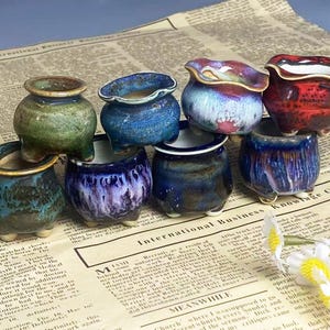 May include: A collection of small, handcrafted ceramic pots in various colors, including green, blue, and red. The pots have unique glaze finishes and sit on a newspaper background. These decorative pots are ideal for small plants.