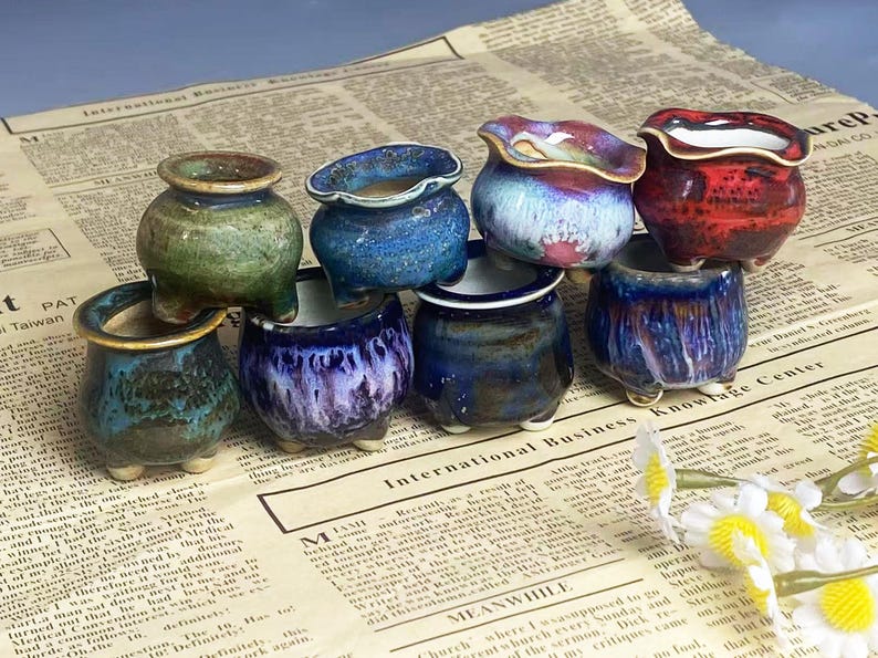 May include: A collection of small, handcrafted ceramic pots in various colors, including green, blue, and red. The pots have unique glaze finishes and sit on a newspaper background. These decorative pots are ideal for small plants.
