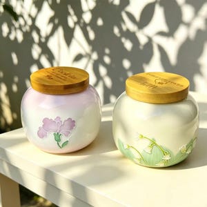 Puede incluir: Dos tarros de cerámica con tapas de madera sobre una mesa blanca. Uno es rosa pálido con un diseño floral, el otro blanco roto con un diseño floral verde. Las tapas son de color marrón claro y tienen texto.