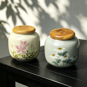 May include: Two ceramic jars with wooden lids. One jar is light green with pink flower illustrations, the other is light blue with white daisy illustrations. The lids have engraved designs. The jars sit on a black table.