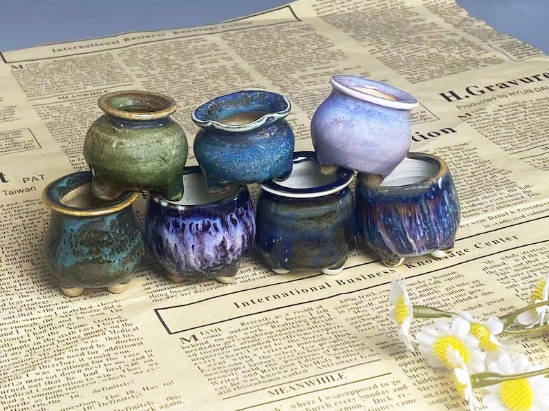 May include: A collection of small, glazed ceramic pots in various colors, including green, blue, and purple. Each pot has a unique shape and sits on three small feet. The pots are arranged on a newspaper background.