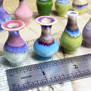 Op de afbeelding: Een verzameling miniatuur keramische vazen met diverse reactieve glazuurafwerkingen in tinten blauw, paars, roze en groen. Een metalen liniaal ligt op de voorgrond en toont aan dat de vazen ongeveer 3,8 cm hoog zijn.