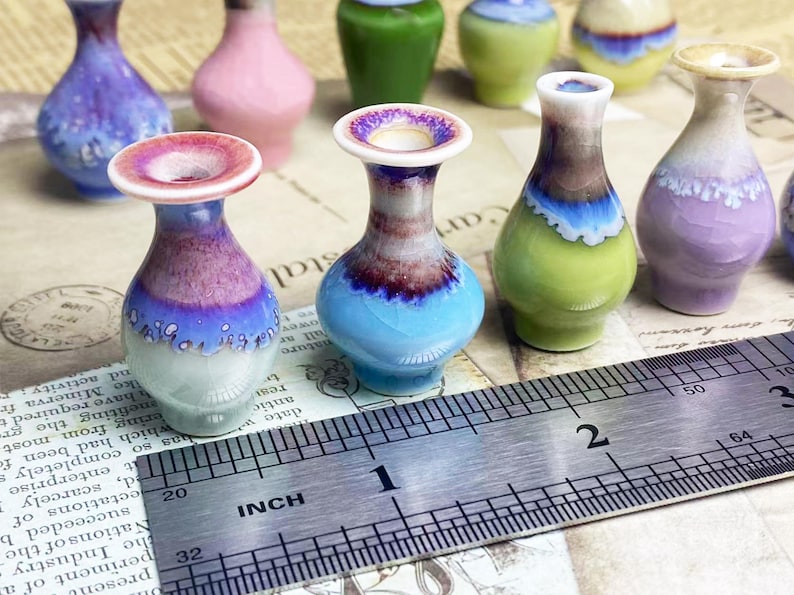 Op de afbeelding: Een verzameling miniatuur keramische vazen met diverse reactieve glazuurafwerkingen in tinten blauw, paars, roze en groen. Een metalen liniaal ligt op de voorgrond en toont aan dat de vazen ongeveer 3,8 cm hoog zijn.