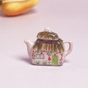 Könnte beinhalten: Kleine rosa Keramik-Teekanne mit Strohdach-Design auf Deckel und Körper. Verziert mit roten Beeren, einer grünen Tür und einem Fenster. Henkel und Ausguss sind ebenfalls rosa.