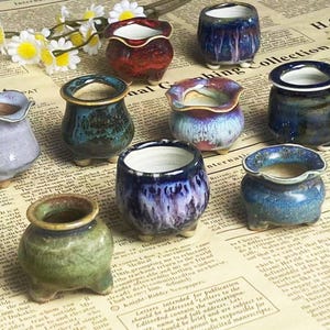 Peut inclure: Ensemble de petits pots en céramique faits à la main, aux couleurs variées, dont le bleu, le vert et le rouge. Chaque pot a un émail unique et repose sur de petits pieds. Les pots sont disposés sur un fond de journal.