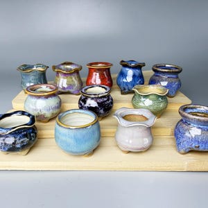 Może przedstawiać: Kolekcja małych, ceramicznych doniczek w różnych kolorach, w tym niebieskim, zielonym, czerwonym i fioletowym. Doniczki mają unikalny, falisty brzeg i są ułożone na drewnianej ekspozycji. Te dekoracyjne doniczki nadają się do małych roślin.