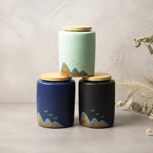 May include: Three cylindrical ceramic canisters with light wooden lids stacked in a pyramid. The top canister is mint green, the bottom left is navy blue, and the bottom right is black. Each features a gold and blue mountain landscape graphic.