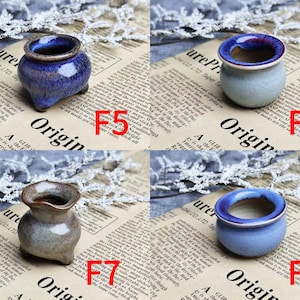 May include: Four small ceramic pots in various colors: blue, gray, and brown. Each pot has a unique shape and design, with a glossy finish. The pots are displayed on a newspaper background with white floral accents.