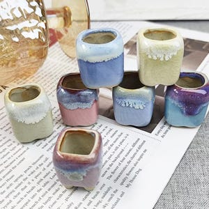 May include: A selection of small, square ceramic plant pots in shades of blue, pink, and green, with a glossy finish. The pots are displayed on a newspaper, suitable for small plants or succulents.