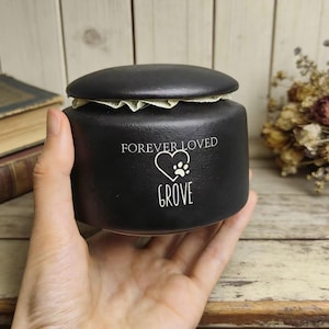 May include: A black pet urn with a lid, held in a hand. The urn has the words "FOREVER LOVED" above a heart and paw print design, with the name "GROVE" below. The background includes books and dried flowers.