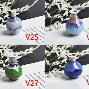 Op de afbeelding: Vier miniatuur keramische vazen gelabeld V25, V26, V27 en V28. V25 is lichtblauw met paarse druppels. V26 is lichtblauw met een paarse en witte hals. V27 heeft een groene basis en een blauw-witte hals. V28 is donkerblauw met een witte hals.