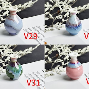Op de afbeelding: Vier miniatuur keramische vazen met glanzende, reactieve glazuren. V29 is lichtblauw en wit, V30 is blauw en bruin, V31 is groen en paars, en V32 is roze en blauw. Elke vaas heeft een smalle hals en een ronde vorm op wit papier.