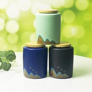 Może przedstawiać: Trzy ceramiczne słoiki w odcieniach niebieskiego, czarnego i miętowego, ułożone na białej powierzchni. Każdy słoik ma drewnianą pokrywkę i namalowany wzór gór i ptaków. Tło jest miękko zielone.