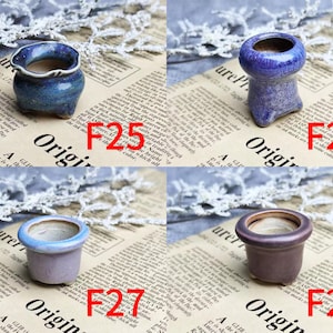 May include: Four small ceramic pots in shades of blue, purple, and brown are displayed. Each pot has a unique shape and design, with a textured or glazed finish. The pots are labeled with the letters "F" and numbers.