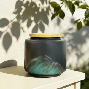 May include: A dark blue ceramic jar with a light wood lid. The jar has a mountain design in shades of green and blue. The jar is sitting on a white surface with a white wall in the background.