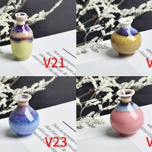 Op de afbeelding: Vier miniatuur keramische vazen gelabeld V21, V22, V23 en V24. V21 is geel met een paarse rand. V22 is beige met een blauwe rand. V23 is blauw met een donkerblauwe rand. V24 is roze met een blauwe rand. Elke vaas heeft een glanzende afwerking.