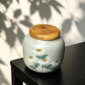 May include: A ceramic jar with a wooden lid. The jar is light green with a floral design of white daisies. The lid is engraved with the text "FOREVER IN OUR HEARTS" and the name "Hillary" with dates.