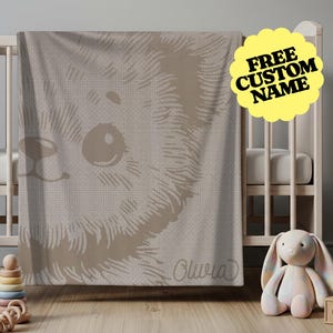 May include: A beige baby blanket with a large, detailed illustration of a bear's face. The blanket is draped over a crib. The text "FREE CUSTOM NAME" is on a yellow, cloud-shaped sticker. The name "Olivia" is printed on the blanket.