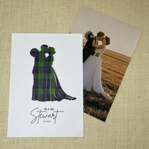 May include: A white rectangular print featuring a silhouette of a couple in a green and blue tartan pattern, with the text "MR & MRS Stewart" and "23.11.2024" below. Beside it lies a photograph of a couple embracing in a field at sunset.