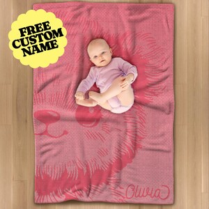 May include: A pink baby blanket with a lion design and the name "Olivia" in cursive. The blanket has a textured appearance and features the text "FREE CUSTOM NAME" in a yellow burst. A baby in a pink onesie is lying on the blanket.