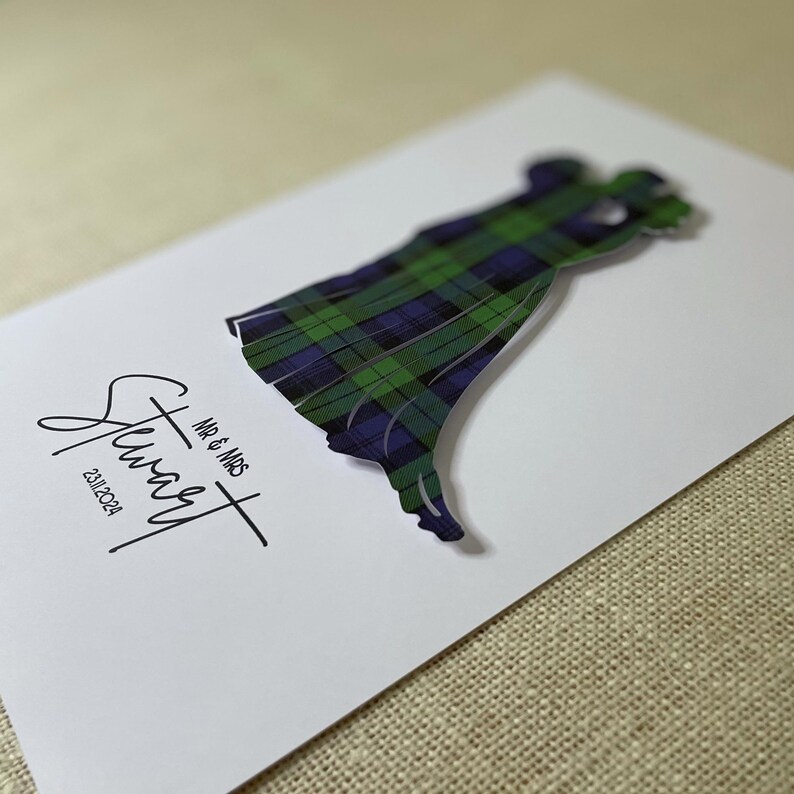May include: A white rectangular card featuring a die-cut silhouette of two figures embracing, filled with a green, blue, and black tartan pattern. The text reads "MR & MRS", "Stuart", and the date "23.11.2024" in black ink on the left side.