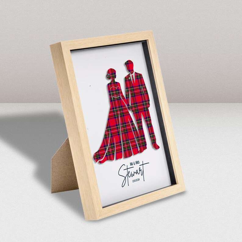May include: A light wood frame containing a white print of two figures in red tartan clothing. The figures represent a couple, with text below reading MR & MRS Stewart and the date 23.8.2024. The frame has a stand and is angled on a surface.