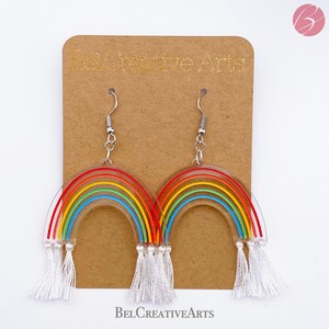 May include: Rainbow-colored acrylic earrings with white tassels. The earrings are shaped like rainbows and have a silver hook.