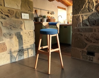 Puede incluir: Un taburete de bar alto de madera con una estructura de madera natural clara, cuatro patas rectas y un respaldo curvo. El asiento y el respaldo están tapizados en una tela azul sólida, lo que crea un contraste con el acabado de madera clara.